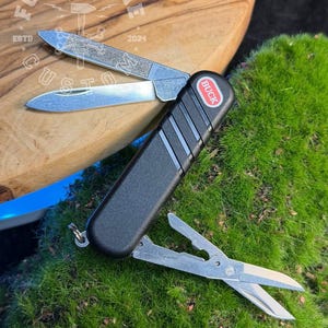 Può includere: Un multiutensile nero con la scritta "BUCK" sul lato, dotato di lima per unghie, due lame e forbici. L'utensile è aperto, mostrando i vari strumenti. Lo sfondo comprende una superficie in legno e muschio verde.