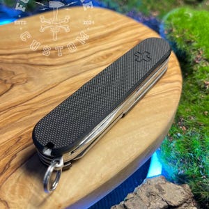 May include: A black and silver multi-tool knife with a textured grip and a silver keyring. The knife features a Swiss cross emblem and multiple tools. The knife is resting on a wooden surface.