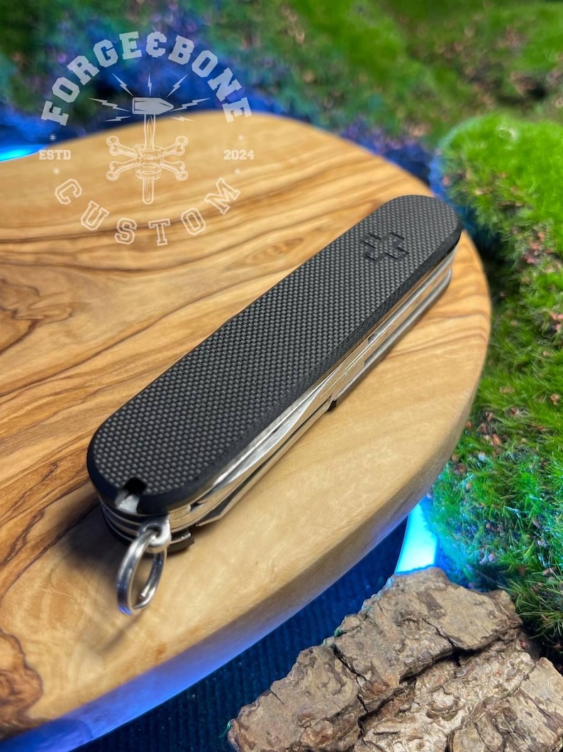 May include: A black and silver multi-tool knife with a textured grip and a silver keyring. The knife features a Swiss cross emblem and multiple tools. The knife is resting on a wooden surface.