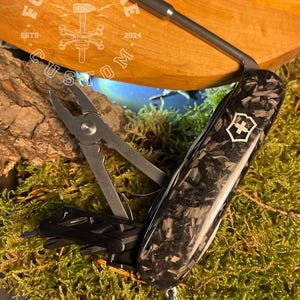 Könnte beinhalten: Ein Multitool mit einem schwarzen Kohlefasergriff und verschiedenen Werkzeugen. Zu den Werkzeugen gehören eine Zange, ein kleines Messer und andere Geräte. Der Text "FORGE&BONE CUSTOM" ist im Hintergrund sichtbar.