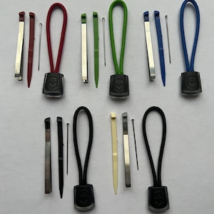 Puede incluir: Seis juegos de tiradores de cremallera con cordones de diferentes colores y un pequeño juego de herramientas. Cada juego incluye una pequeña herramienta de metal, un pequeño alfiler de metal y una pequeña herramienta de plástico. Los tiradores de cremallera son negros con un logotipo blanco.