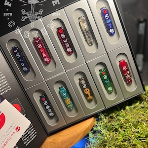 Könnte beinhalten: Ein Set mit zehn individuellen Taschenmessern in einem schwarzen Präsentationskoffer. Die Messer haben verschiedene Designs und Farben, darunter Rot, Blau, Grün, Schwarz und Weiß. Der Koffer trägt die Aufschriften "FORGE & BONE" und "CUSTOM".