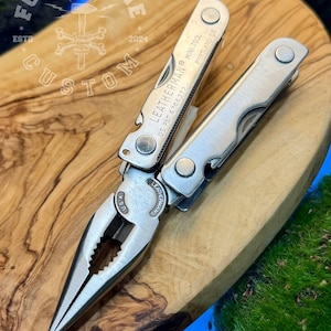 Könnte beinhalten: Ein silbernes Leatherman Mini Tool Multitool mit ausgezogener Zange. Das Werkzeug ist mit "LEATHERMAN MINI TOOL PORTLAND OR" und "US PAT 4744272" gekennzeichnet. Das Werkzeug liegt auf einer Holzoberfläche mit dem Text "FORGE & BONE CUSTOM" und "ESTD 2024".