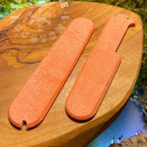 May include: Two orange-coloured, textured, rectangular tools with rounded edges. The tools are on a wooden surface. The text "FORGECBONE ESTD 2024 CUSTOM" is visible on the surface.