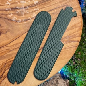 May include: Two dark green textured knife handle scales on a wooden surface. One scale has a cross emblem, and the other has a notch. The wooden surface has the text "FORGEBONE CUSTOM" and a logo.