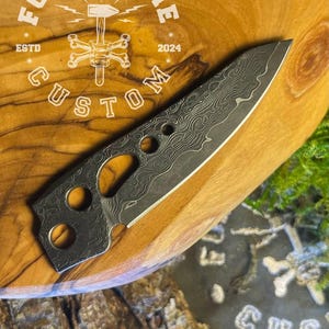 May include: A close-up of a custom-made knife with a damascus steel blade and a unique design. The knife has several holes in the blade and is resting on a wooden surface. The knife is made by Forge & Bone Custom, established in 2024.