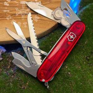 May include: A red Swiss Army knife with multiple tools extended, including a blade, scissors, saw, and screwdriver. The knife features the Victorinox brand name and a white cross emblem. The knife is on a wooden surface with the text "FORGECBON CUSTOMS ESTD".