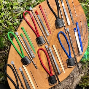 May include: A selection of multi-coloured lanyard tools and accessories, including red, green, blue, and black lanyards with matching tools. The tools are arranged on a wooden cutting board.
