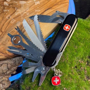 Può includere: Un multiutensile nero con vari strumenti, tra cui un coltello, una sega e una lima. Presenta un emblema a croce rosso e bianco e un piccolo ciondolo rosso con una croce. Lo strumento è esposto su una superficie di legno.