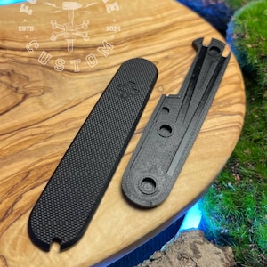 May include: Two black custom parts, one rectangular with a textured surface and the other with a unique shape. The parts are displayed on a wooden surface with the "Forge & Bone Custom" logo. The logo includes the text "ESTD 2024".