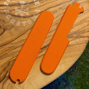 Puede incluir: Dos cachas de mango de cuchillo de color naranja brillante y texturizadas sobre una tabla de cortar de madera. Las cachas tienen forma rectangular con extremos redondeados y una superficie texturizada. El logotipo "FORGE & BONE CUSTOM" es visible.