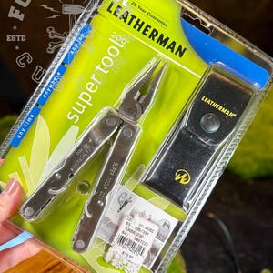 以下が含まれることがあります： シルバーメタルのレザーマン スーパーツール200マルチツールと黒いレザーシースが、ライムグリーンとブルーのブリスターパックに入っています。パッケージには「Leatherman」「Super Tool 200」「25 Year Guarantee」の文字と79.99ドルの価格タグが記載されています。