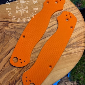 Può includere: Due guancette per manico di coltello arancioni brillanti con una superficie strutturata e fori multipli per viti. Le guancette sono curve e progettate per adattarsi a un coltello. L'immagine include il testo "FORGE & BONE CUSTOM ESTD 2024".