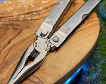 Könnte beinhalten: Ein silbernes Multitool mit Zange, Messer und anderen Werkzeugen, das auf einer Holzoberfläche liegt. Das Werkzeug ist mit dem Text "LEATHERMAN WAVE" und "US REG TM D335473" graviert. Das Bild enthält auch den Text "FORGE&BONE CUSTOM ESTD 2024".