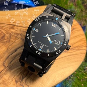 May include: Black wristwatch with a black dial, blue accents, and a date display. The watch features the text "SWISS MOVEMENT 200M" and "LEATHERMAN". The watch is on a wooden surface with the text "FORGE & BONE CUSTOM" and the year "2024".