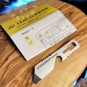 Könnte beinhalten: Ein Leatherman Brewzer Multitool und eine Bedienungsanleitung werden auf einer Holzoberfläche gezeigt. Der Brewzer ist silberfarben mit einer schwarzen Kante und dem Leatherman-Logo. Die Anleitung ist gelb und weiß mit dem Text "Piranha - Mako - Ti Brewzer User Guide."