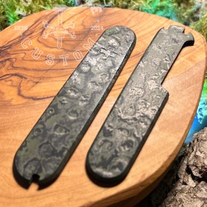 Puede incluir: Dos herramientas de metal personalizadas con un patrón de camuflaje en tonos verdes y grises. Una tiene un símbolo de cruz, la otra un extremo dentado. Se muestran sobre una superficie de madera con el texto "FORGE CUSTOM" y el año "2024".