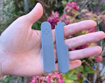 May include: Two grey plastic scales for a pocket knife. The scales have a cross embossed on one side and a smooth surface on the other.