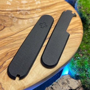 May include: Two black, textured accessories with a carbon fibre pattern are displayed on a wooden surface. One is rectangular, the other has a curved end and a notch. The wooden surface has the text "FORGE & BONE CUSTOM" and the year "2024".