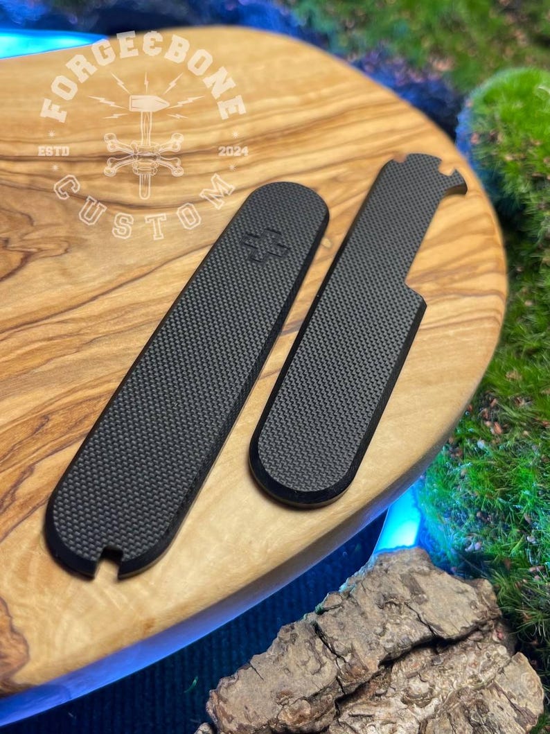 May include: Two black, textured accessories with a carbon fibre pattern are displayed on a wooden surface. One is rectangular, the other has a curved end and a notch. The wooden surface has the text "FORGE & BONE CUSTOM" and the year "2024".