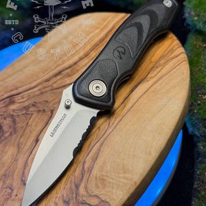 Puede incluir: Una navaja plegable con una hoja parcialmente dentada de color plateado marcada con la palabra LEATHERMAN. El mango es negro con un agarre texturizado y un tornillo de pivote plateado. El logotipo FORGE&BONE CUSTOM ESTD 2024 es visible.