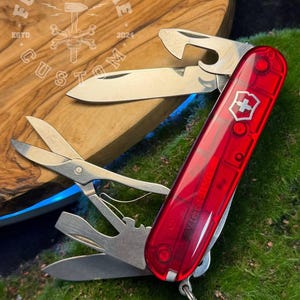 May include: A red Swiss Army knife with various tools, including a blade, scissors, and bottle opener. The knife has the Victorinox logo and is set against a wooden surface and green grass.