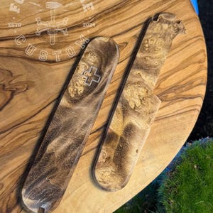 Puede incluir: Dos artículos de madera personalizados con un patrón de vetas naturales. Uno es rectangular con un diseño de cruz, el otro tiene forma de cuchillo. Están sobre una superficie de madera con el texto "FORGE & BONE CUSTOM" y el año "2024".
