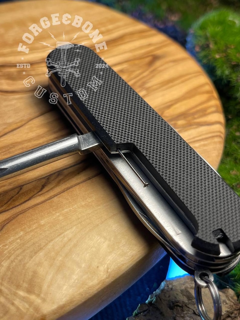 May include: A multi-tool with a textured black handle and a silver metal body. The tool includes various implements and a key ring attachment. The text "FORGE & BONE CUSTOM" is printed on the handle.