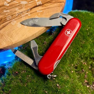 Könnte beinhalten: Ein rotes Schweizer Taschenmesser mit mehreren Werkzeugen, darunter eine Klinge, eine Nagelfeile und ein Flaschenöffner. Das Messer hat ein weißes Kreuzemblem und liegt auf grünem Moos und einer Holzoberfläche mit dem Text "FORGE&BONE CUSTOM".