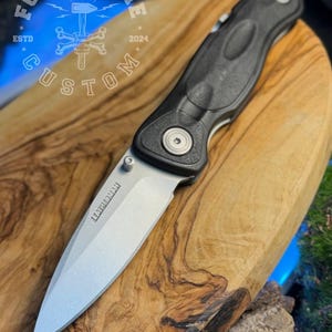 以下が含まれることがあります： シルバーの金属製ブレードと黒いテクスチャ加工されたハンドルを備えたレザーマンの折りたたみナイフ。ブレードの表面にはブランド名「LEATHERMAN」が刻印されています。「FORGEBONE CUSTOM ESTD 2024」というテキストが記された円形のロゴが確認できます。