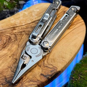 May include: A multi-tool with pliers, knife, and other tools, made of stainless steel. The tool is open, displaying the pliers. The brand name "LEATHERMAN FREE P4" is visible. The tool is on a wooden surface. The text "FORGE & BONE CUSTOM" is in the background.