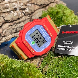 Könnte beinhalten: Eine rote und goldene G-Shock-Uhr mit einem Super Mario Bros.-Design. Das Zifferblatt der Uhr zeigt die Zeit, das Datum und den Wochentag an. Das Armband der Uhr ist aus rotem und goldenem Harz. Die Uhr liegt auf einem Bett aus grünem Moos. Der Text auf der Uhr lautet "Super Mario Bros. Casio" und "G-Shock".