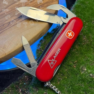 Può includere: Un coltellino svizzero multifunzione rosso con un emblema a croce bianca e la parola LASER stampata sopra un'icona triangolare a raggiera. L'utensile presenta una lama principale, un apribottiglie e una lima per unghie, con un anello metallico.