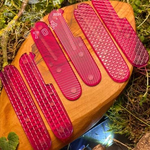 Puede incluir: Seis escamas de plástico rosa para una navaja de bolsillo, con diferentes patrones, sobre una superficie de madera. Las escamas están dispuestas en fila, con la más grande en la parte superior y la más pequeña en la parte inferior. Las escamas están todas hechas de un plástico translúcido, y tienen una variedad de diferentes patrones. Las escamas están todas enfocadas, y el fondo está desenfocado.