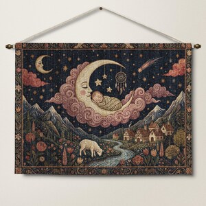 May include: A rectangular wall tapestry featuring a woven scene of a swaddled baby sleeping on a crescent moon surrounded by pink clouds, stars, and a dreamcatcher. Below, a white sheep grazes in a meadow near a winding river and a small village.