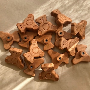 以下が含まれることがあります： それぞれユニークなデザインと中央に小さな穴のある、木製のクマの形をしたおもちゃのコレクション。おもちゃは天然木で作られており、薄い茶色をしています。
