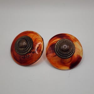 May include: A pair of vintage clip-on earrings. Each earring features a round, marbled orange and brown design with a bronze-colored center and a twisted rope border. The earrings have a retro aesthetic.