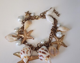 May include: A gold-toned chain bracelet adorned with seashells, faux pearls, and starfish charms. The bracelet features a variety of seashells in shades of white and brown, with faux pearls interspersed throughout the design. The starfish charms add a nautical touch.