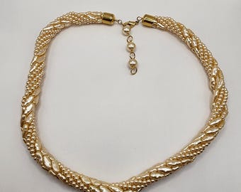 May include: A cream-colored necklace with a braided design. The necklace features small, round beads and larger, leaf-shaped beads. The clasp and extender chain are gold-toned, and the necklace is set against a white background.