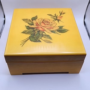 May include: A yellow wooden box with a floral design on the lid. The design features a pink and white rose with green leaves.