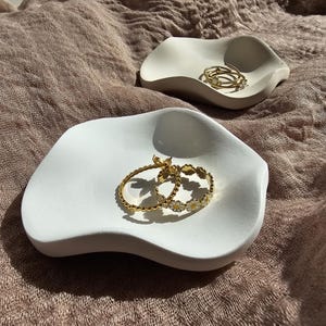 Könnte beinhalten: Zwei weiße, organisch geformte Ringschalen auf einem strukturierten, beigefarbenen Stoff. Eine Schale enthält mehrere goldene Ringe, darunter einen Daisy-Ring. Die andere Schale enthält mehrere goldene Ringe. Die Schalen sind schlicht und elegant.