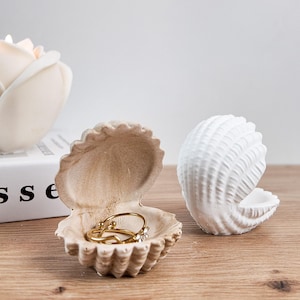 Könnte beinhalten: Zwei muschelförmige Ringhalter. Einer ist offen und zeigt goldene Ringe mit Blumenakzent. Der andere ist geschlossen und weiß. Beide stehen auf einer Holzoberfläche. Eine weiße Blume und ein Buch sind im Hintergrund.