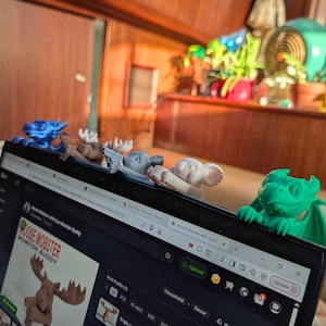 May include: A laptop displays a product page for a "Moose Mobster Monitor Buddy." Various 3D-printed figurines, including a moose, koala, and dragon, are perched on the laptop's top edge. The figurines are in shades of blue, gray, and green.
