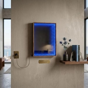 Puede incluir: Un espejo infinito con marco de madera y borde de luz LED azul está montado en una pared texturizada. Una pequeña placa de latón debajo del espejo dice "INFINITY MIRROR 25X38CM". A la derecha, una estantería con libros y un jarrón con flores.