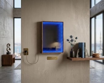 Könnte beinhalten: Ein Infinity-Spiegel mit Holzrahmen und blauem LED-Lichtrand ist an einer strukturierten Wand montiert. Ein kleines Messingschild unter dem Spiegel lautet "INFINITY MIRROR 25X38CM". Rechts befindet sich ein Regal mit Büchern und einer Blumenvase.