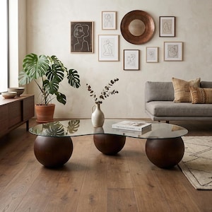 Puede incluir: Un salón moderno con una mesa de centro con tapa de cristal sostenida por tres esferas de madera oscura. La habitación incluye un sofá gris, dibujos lineales enmarcados, una gran planta en maceta y cojines decorativos. Un espejo circular de color cobre cuelga en la pared.
