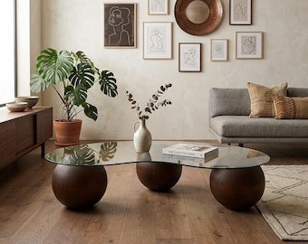May include: A modern living room scene featuring a glass-topped coffee table with three dark wooden spherical bases. The room includes a gray sofa, framed line art, a large potted plant, and decorative pillows. A copper-colored circular mirror hangs on the wall.