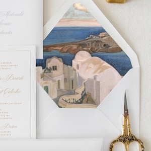 May include: A white envelope with a gold border, featuring a watercolor painting of a white building with a blue roof, overlooking a blue body of water. The envelope is partially open, revealing a white card with gold lettering.