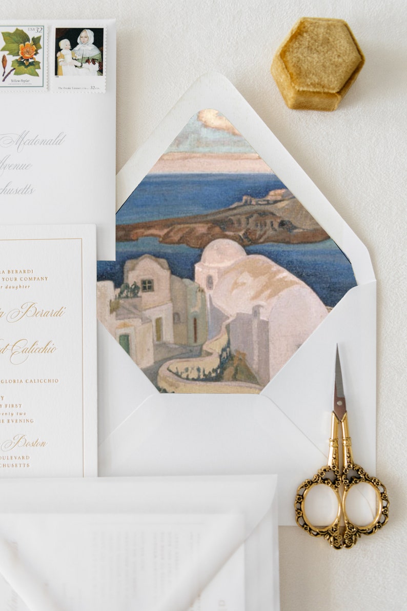 May include: A white envelope with a gold border, featuring a watercolor painting of a white building with a blue roof, overlooking a blue body of water. The envelope is partially open, revealing a white card with gold lettering.