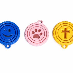May include: Three colorful pet tags: a blue tag with a winking smiley face, a pink tag with a paw print, and a yellow tag with a cross. Each tag has a circular design with a textured edge and a hole for attachment.